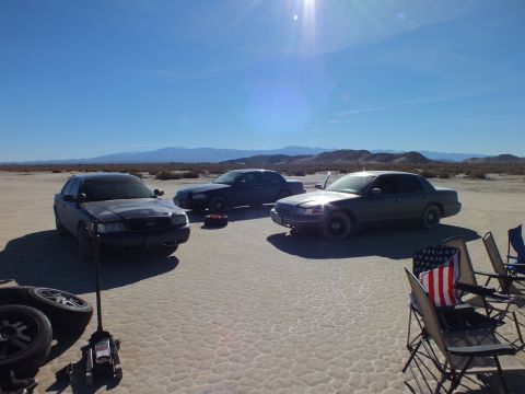 2003 Ford Crown Victoria - Sedans parked on cracked desert flats facing distant mountains, lawn chairs and US flag at the edge