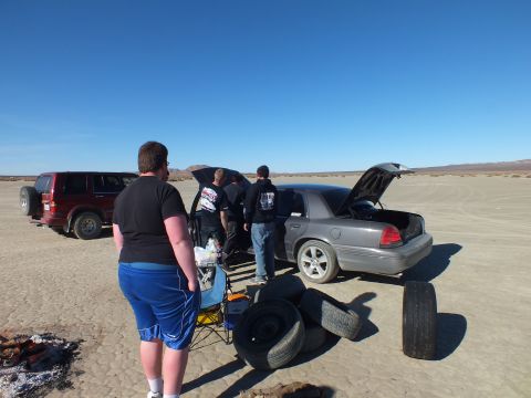 2003 Ford Crown Victoria - Onlookers around a silver Ford sedan with hood and trunk open, used tires and an SUV on the lakebed