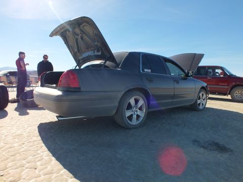 2003 Ford Crown Victoria - Rear view of a gray full-size sedan with trunk up, people and a red SUV behind on the lakebed