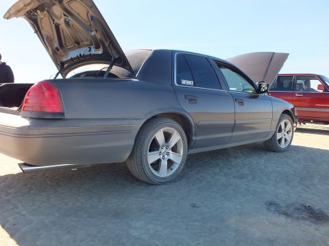 2003 Ford Crown Victoria - Side-rear of a gray full-size sedan on the lakebed, hood and trunk raised, red vehicle in background