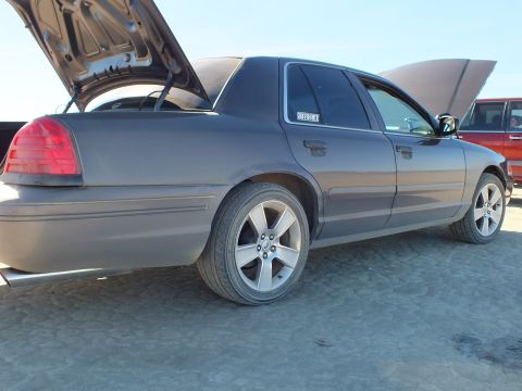 2003 Ford Crown Victoria - Rear quarter of a gray full-size Ford sedan with raised hood and trunk on cracked desert ground