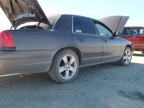 2003 Ford Crown Victoria - Side-rear of a gray full-size sedan on the lakebed, hood and trunk raised, red SUV behind
