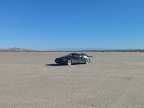 2003 Ford Crown Victoria - Lone dark sedan on cracked desert flats beneath a wide blue sky and far mountain range