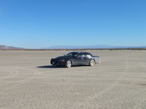 2003 Ford Crown Victoria - Black sedan sits on cracked desert flats with a distant snow-dusted mountain range