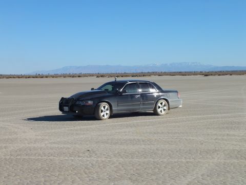 2003 Ford Crown Victoria - Black full-size Mercury sedan parked on cracked desert lakebed before a far mountain range