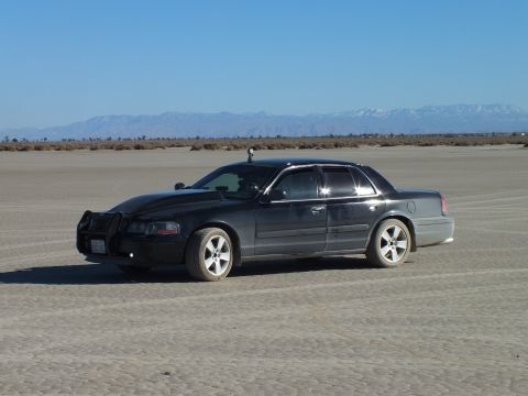 2003 Ford Crown Victoria - Side profile of a black full-size Mercury sedan on cracked desert flats near distant peaks