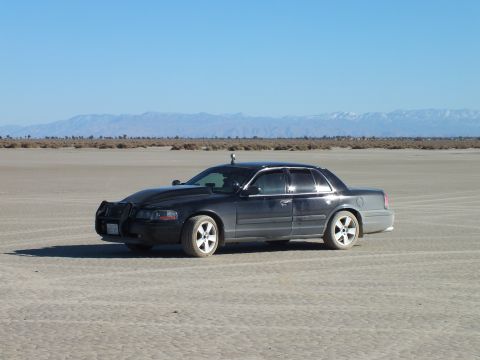 2003 Ford Crown Victoria - Black full-size Mercury sedan parked on cracked desert flats before a far mountain range