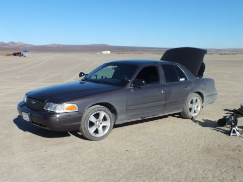 2003 Ford Crown Victoria - Front three-quarter of a gray full-size Ford sedan with trunk up on the lakebed, tripod nearby