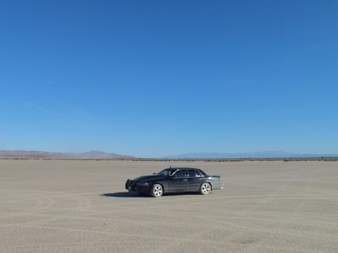 2003 Ford Crown Victoria - Dark full-size sedan alone on cracked desert flats with distant hills and faint dust trail