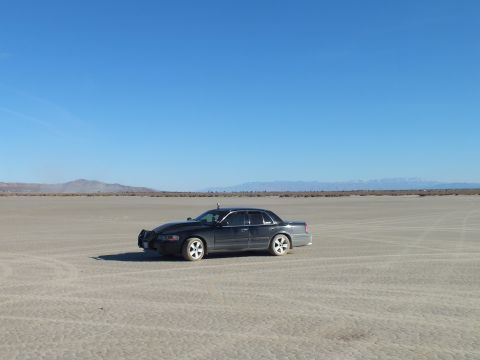 2003 Ford Crown Victoria - Black full-size sedan on cracked desert flats before a distant snow-dusted mountain range