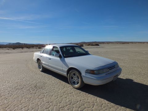 2003 Ford Crown Victoria - Front three-quarter of a white full-size Ford sedan on cracked desert flats under bright sun
