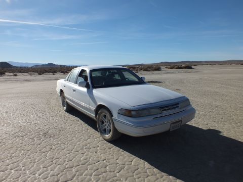 2003 Ford Crown Victoria - Front three-quarter of a white full-size Ford sedan on cracked desert flats under blue sky