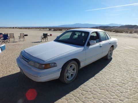 2003 Ford Crown Victoria - Front three-quarter view of a white full-size Ford sedan on cracked desert flats, mountains on the horizon.