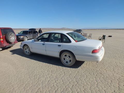2003 Ford Crown Victoria - White full-size sedan parked next to a maroon SUV on cracked desert ground, mountains beyond.