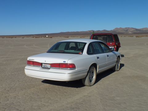 2003 Ford Crown Victoria - White Ford sedan rear quarter on cracked desert ground with a red SUV alongside.