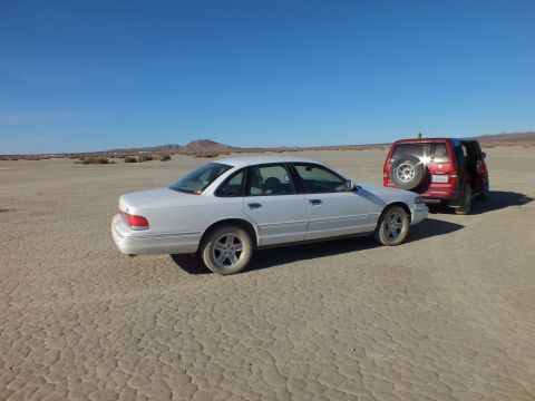 2003 Ford Crown Victoria - Older silver Ford sedan with alloy wheels parked on cracked playa next to a maroon SUV.