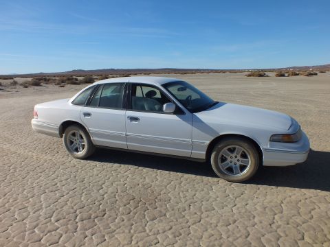 2003 Ford Crown Victoria - Full passenger-side view of a white Ford sedan on dry playa, brush and hills on the horizon.