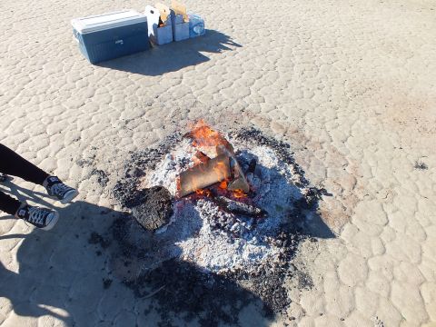 2003 Ford Crown Victoria - Small wood fire and ash ring on cracked playa with coolers and someone standing close by.