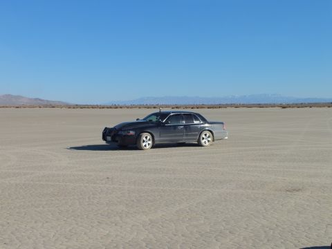 2003 Ford Crown Victoria - Black full-size sedan with mesh grille sits on open cracked playa, snowy mountains far behind.