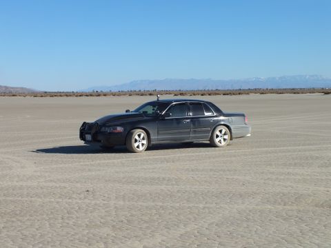 2003 Ford Crown Victoria - Side view of a dark full-size sedan on cracked playa, snow-capped range on the horizon.