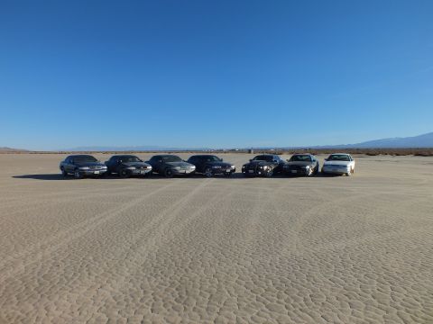 2003 Ford Crown Victoria - Seven sedans parked side by side facing forward on cracked playa under clear blue sky.