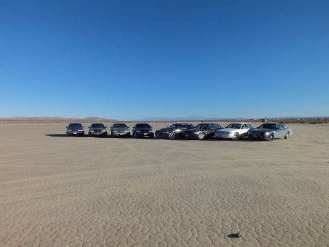 2003 Ford Crown Victoria - Angled lineup of eight sedans on cracked playa with low desert hills in the distance.