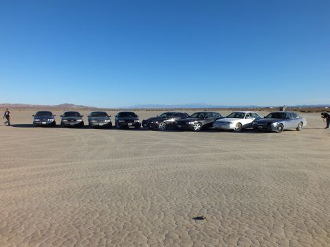 2003 Ford Crown Victoria - Row of eight full-size sedans on cracked playa with people framing the lineup.