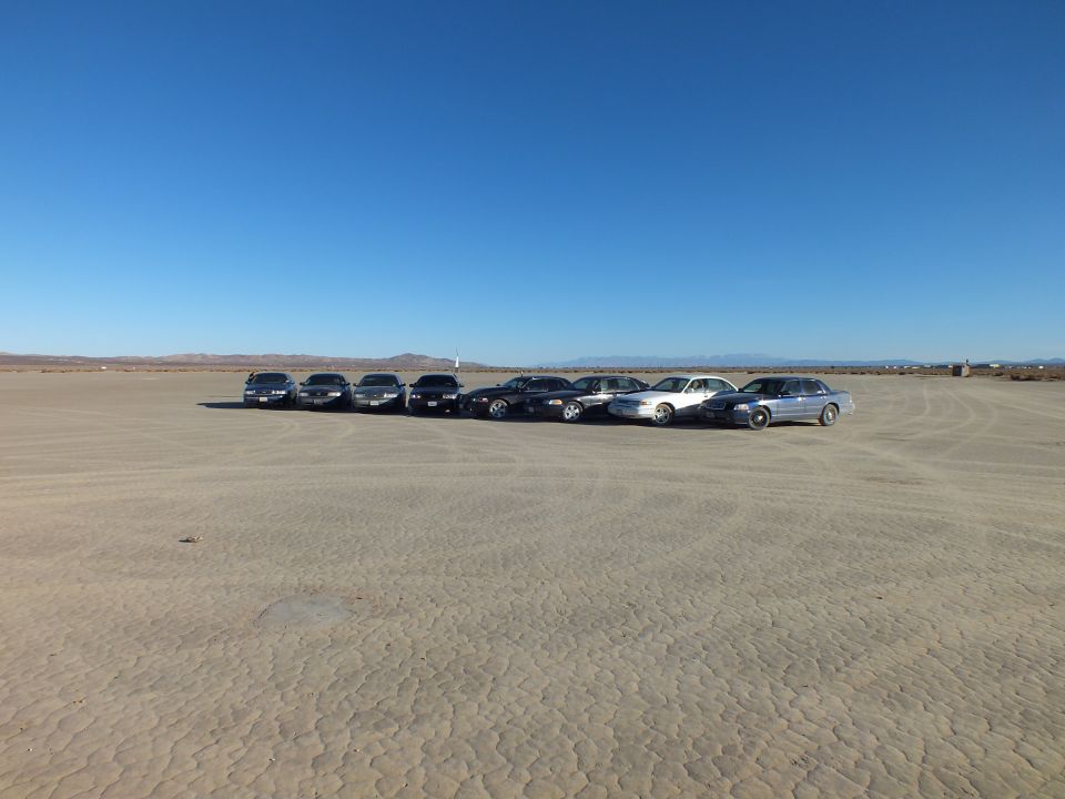 Eight full-size sedans angled across a desert dry lakebed under a clear blue sky