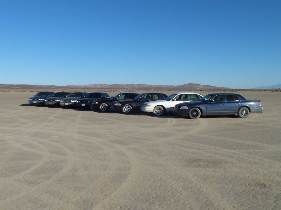 Line of dark sedans and a silver car parked on cracked desert ground