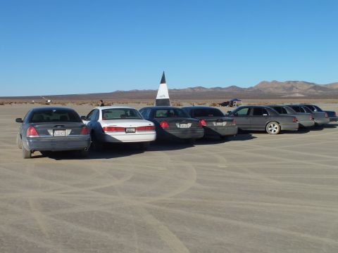 2003 Ford Crown Victoria - Vehicles lined up on playa beside a tall pointed marker and mountains