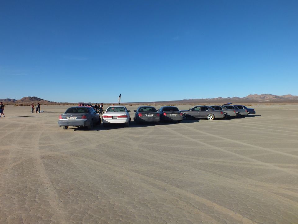 Line of sedans and wagons on dry lakebed with people standing nearby
