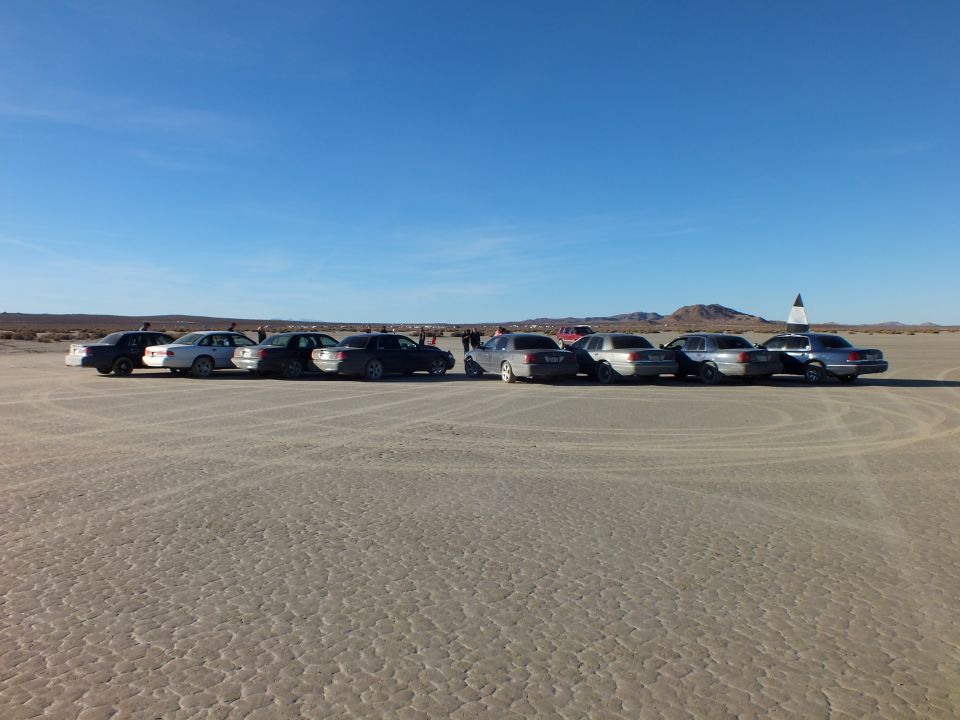 Long row of sedans and wagons on dry lakebed with people and red SUV