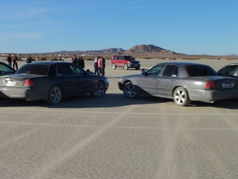2003 Ford Crown Victoria - Black and gray full-size sedans facing each other on a dry lakebed