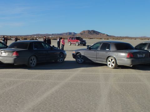 2003 Ford Crown Victoria - Black and silver sedans nose to nose on playa with onlookers and red SUV