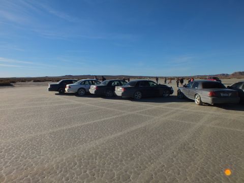 2003 Ford Crown Victoria - Sedans lined up side by side on cracked desert playa with people gathered near distant hills.