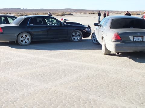2003 Ford Crown Victoria - Two Panther-platform Fords on desert flat; rear plate reads 5VWN8FJ with people in background.