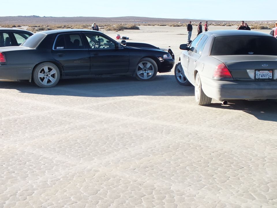 Black Ford Crown Victoria and silver sedan parked on dry lakebed, California plate 5VWN8FJ