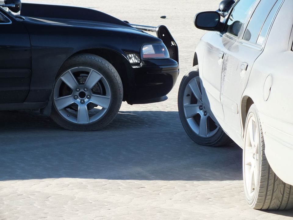 Close-up of black sedan front end beside white sedan, both with five-spoke alloy wheels on dirt