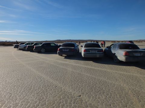 2003 Ford Crown Victoria - Diagonal row of full-size sedans on cracked playa, tire tracks leading toward distant ridgeline.