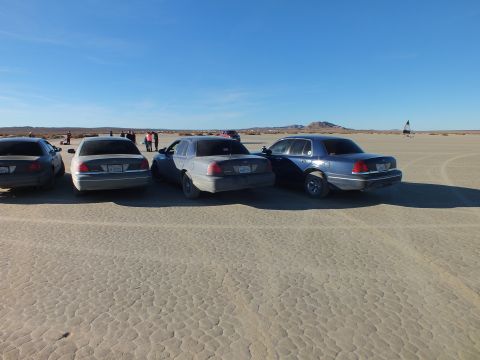 2003 Ford Crown Victoria - Backs of full-size Ford sedans on a dry flat; blue Crown Victoria at right, flag and hills beyond.