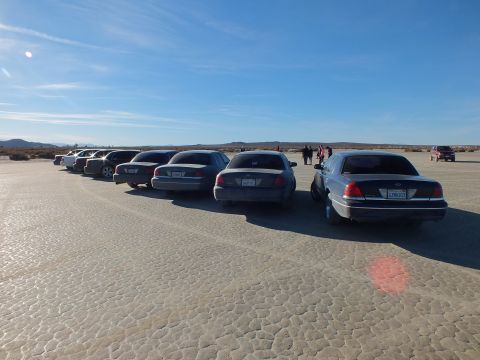 2003 Ford Crown Victoria - Many full-size Ford sedans staggered in a line on cracked playa with mountains on the horizon.
