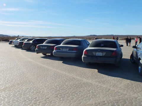 2003 Ford Crown Victoria - Taillights and trunks of full-size Fords lined across packed desert ground, people standing at right.