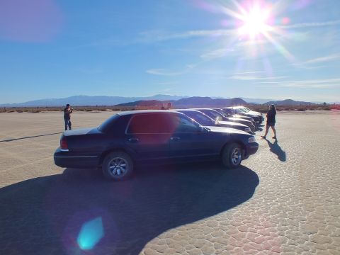 2003 Ford Crown Victoria - Side profile of a navy Panther sedan heading a parked line on desert flat under strong sun flare.