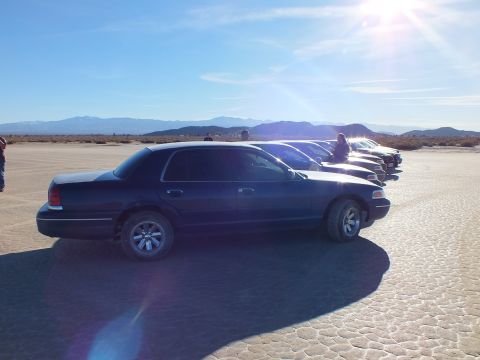 2003 Ford Crown Victoria - Navy full-size Ford shown side-on at the front of a parked line, mountains and sun flare behind.