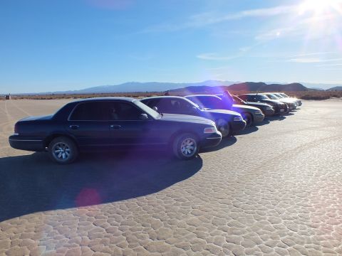 2003 Ford Crown Victoria - Side view of staggered Panther sedans on cracked playa, backlit by low sun over distant peaks.