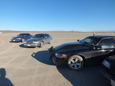 2003 Ford Crown Victoria - Dark Panther sedan close in frame on desert flat with two more Fords parked in the distance.