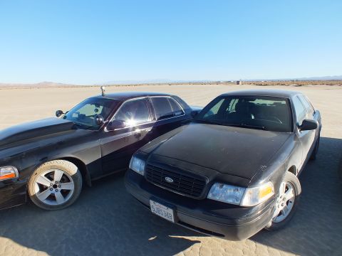 2003 Ford Crown Victoria - Pair of dark Panther Fords on cracked playa; front car's California plate reads 5JD534G.