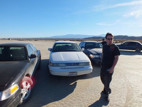 2003 Ford Crown Victoria - Person stands next to a white Panther sedan among parked Fords on desert flat; plate 2NSR413.