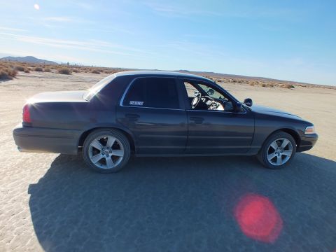 2003 Ford Crown Victoria - Lowered dark Panther sedan shown side-on across cracked desert ground under blue sky.