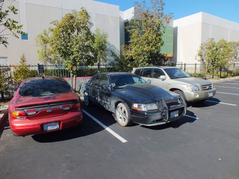 2003 Ford Crown Victoria - Matte-black Panther sedan with grille guard between a red Ford coupe and a silver SUV in a lot.